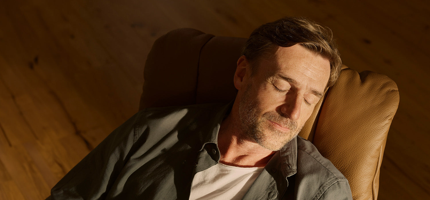 Man resting with eyes closed on a brown leather chair in a warm-toned room.